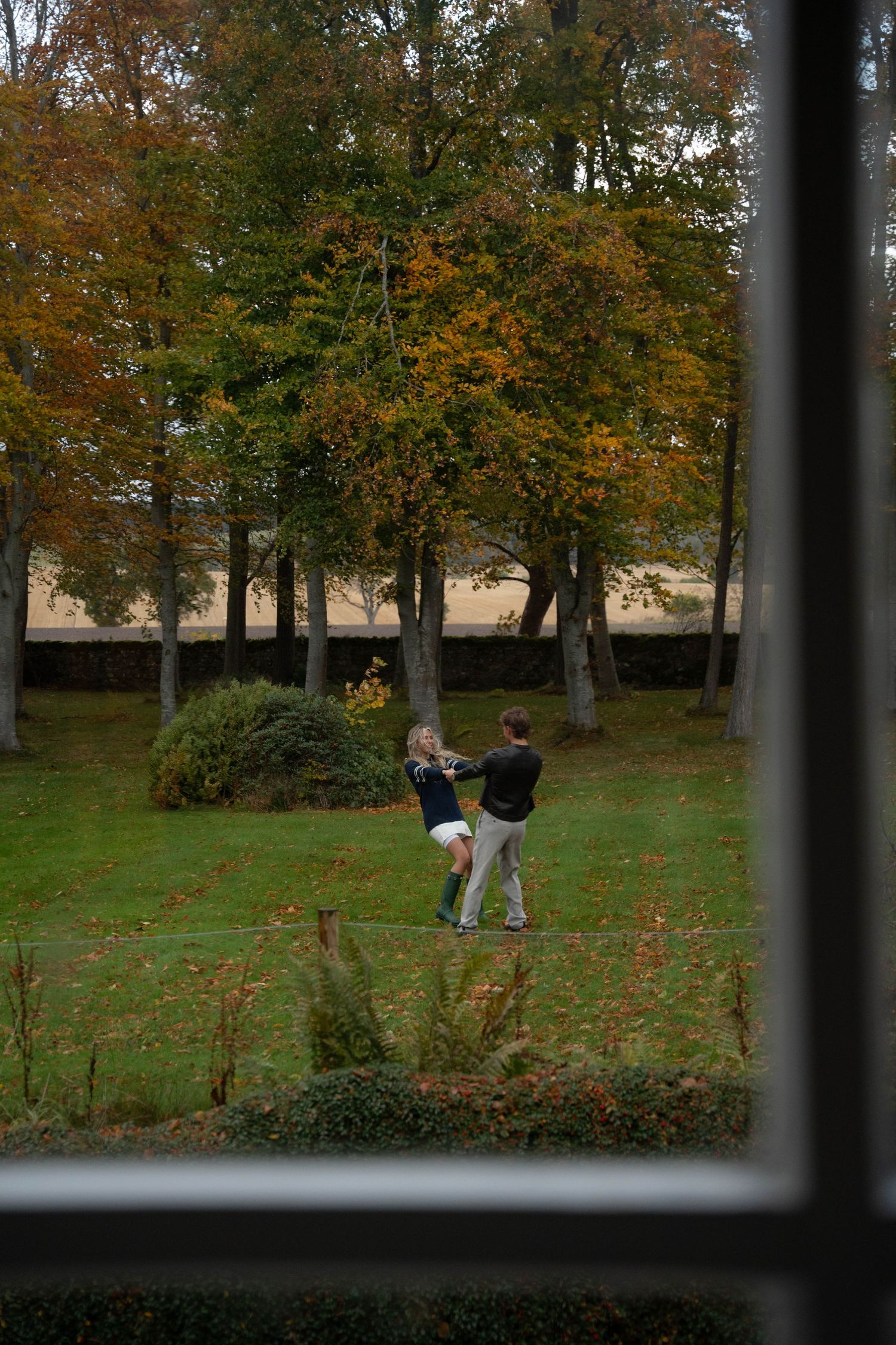 Couple dancing and laughing through a window in Scotland, surrounded by warm fall vibes, capturing a playful and romantic moment.