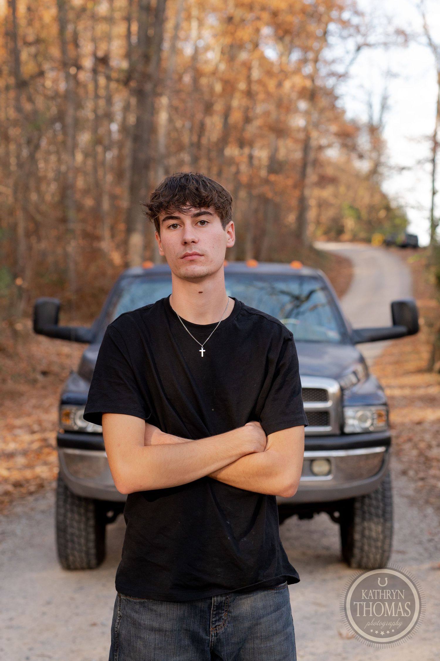 Person in black t-shirt posing in front of lifted Dodge truck with fall foliage in background.