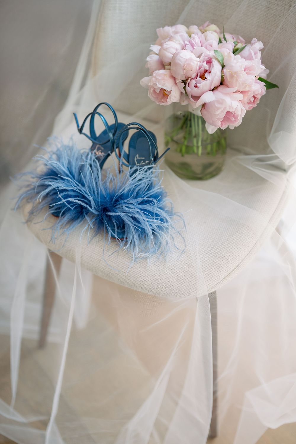 Light pink peonies in a glass vase next to blue feathered fascinator on white tulle fabric.