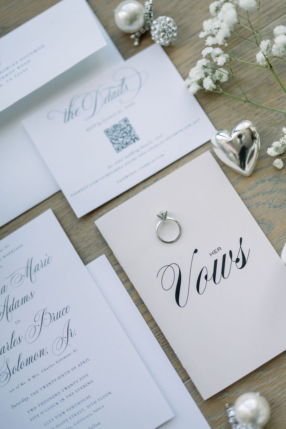 Elegant wedding invitation suite with vow book, rings, and baby's breath flowers on wooden surface.