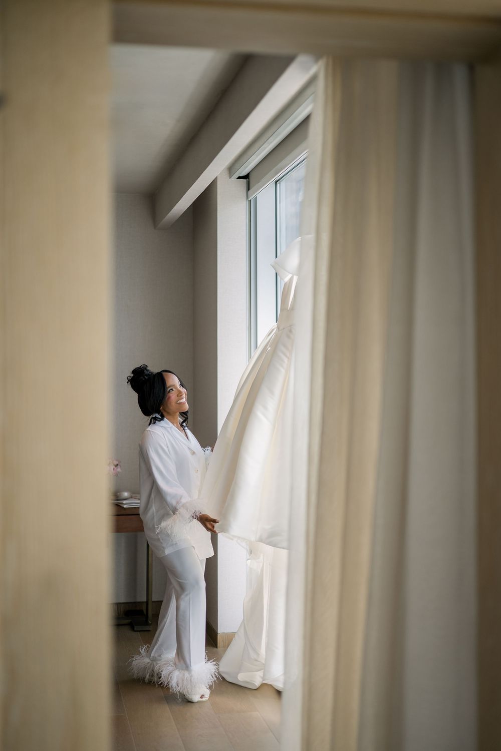 A person in white attire adjusts curtains by a tall window in a bright minimal interior setting.