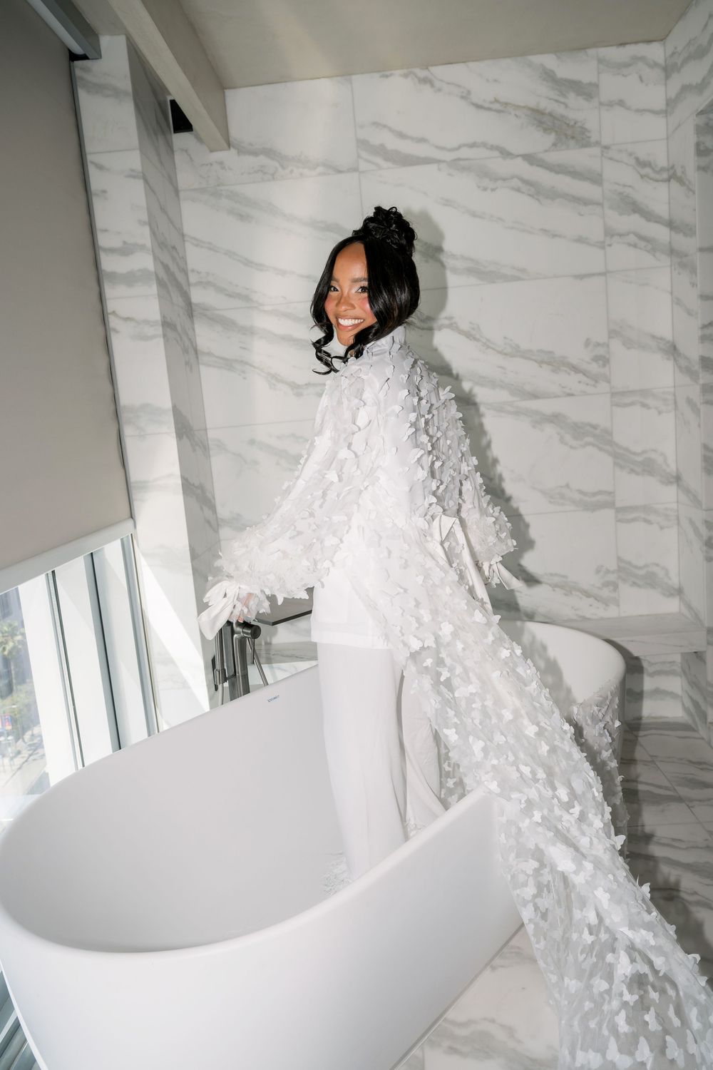 A figure in white poses with a flowing veil near a freestanding bathtub in a luxurious marble bathroom.
