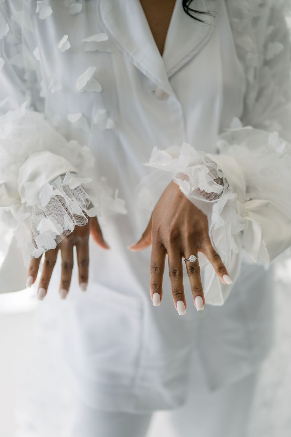 Close-up of hands with white lace gloves and engagement ring against white fabric.