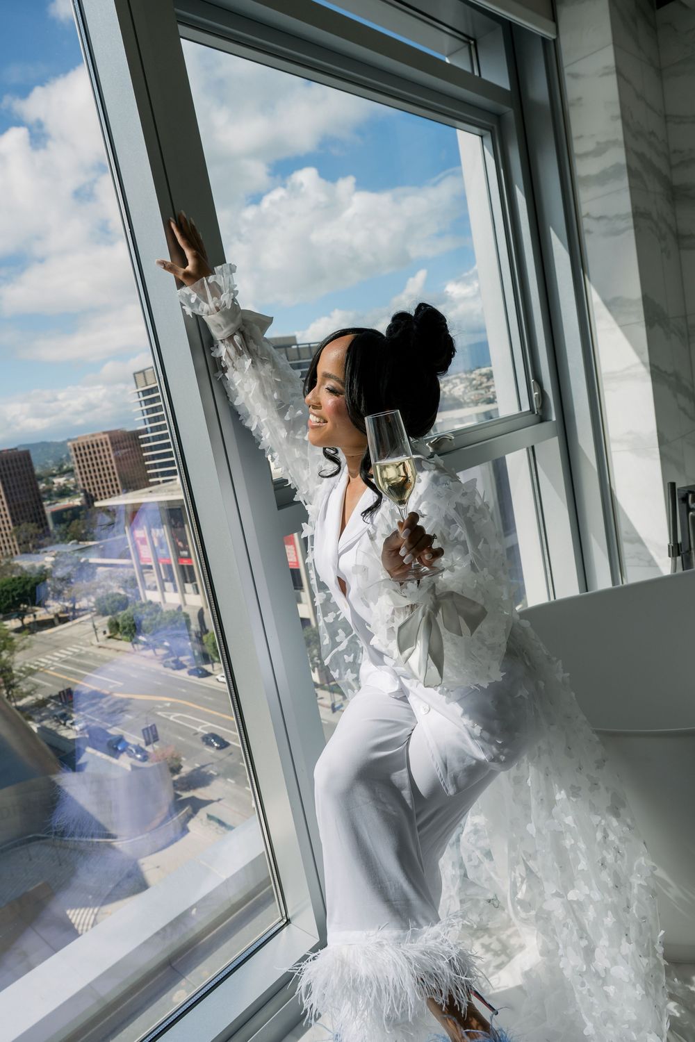 Woman in elegant white outfit standing by window with cityscape view and dramatic light.