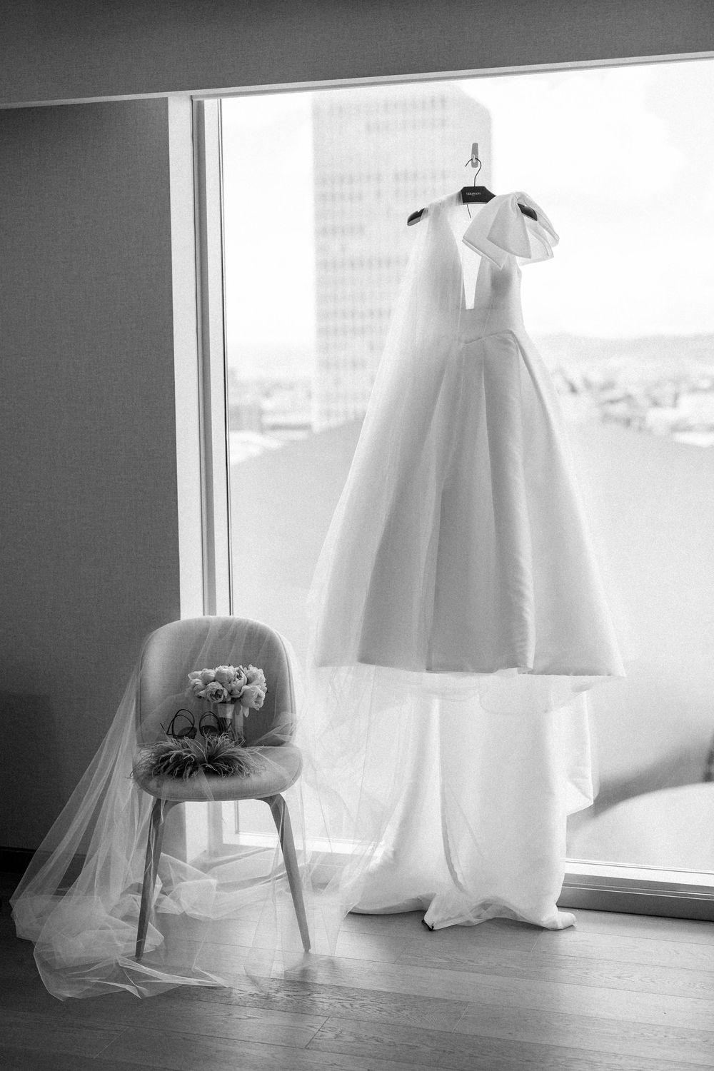 A white wedding dress hangs elegantly by a window with a veil draped over a chair in a black and white photograph.