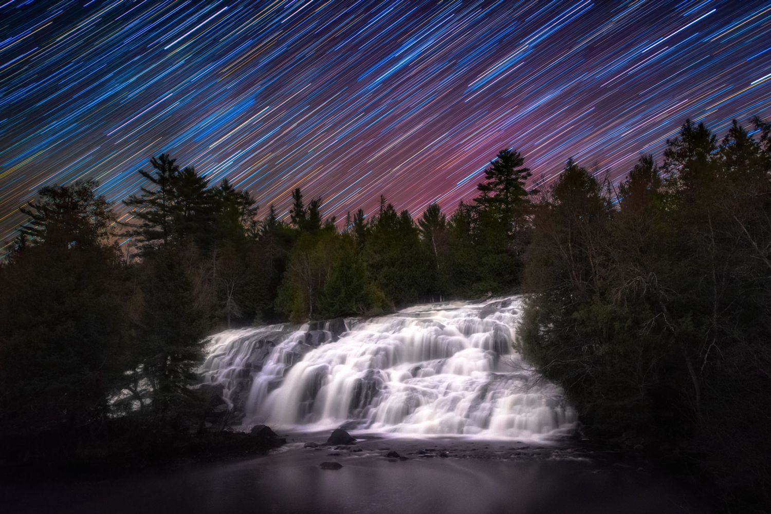 Long exposure photo of a waterfall at night with star trails creating colorful streaks across the dark blue sky.