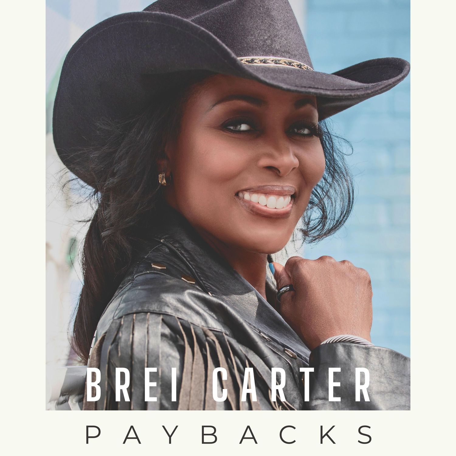 Country artist Brei Carter wearing black cowboy hat and leather jacket poses against blue background for album cover for "Paybacks".