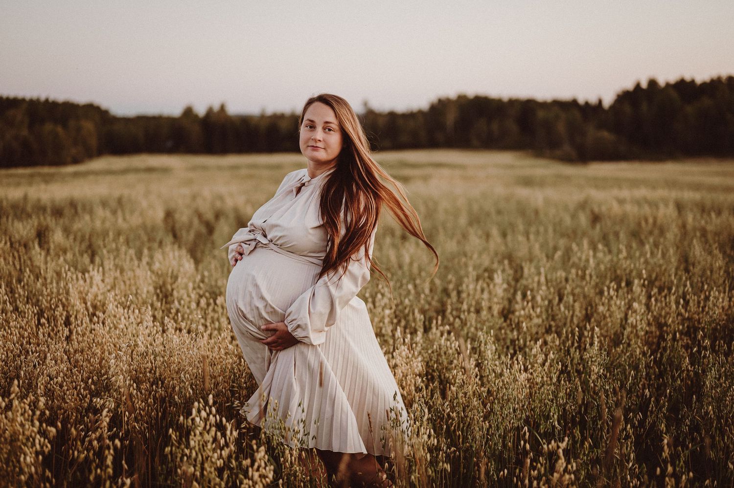 Future maman en robe blanche tournoyant dans un champ de blé doré au coucher du soleil.