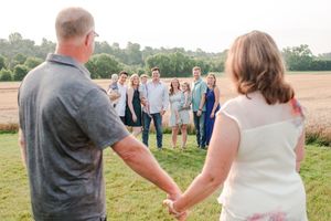Couple holds hands facing large family group gathered in golden field at sunset for outdoor family portrait.