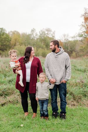Family of four with toddlers standing in autumn meadow, parents in red cardigan and gray hoodie.