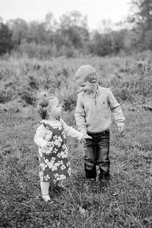Black and white photo of young boy and toddler girl holding hands in grassy outdoor field.