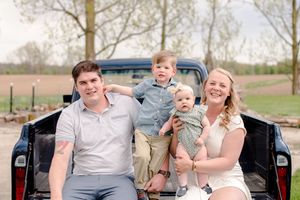 Young family of four poses on vintage truck tailgate in rural setting during spring family photo session.