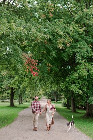 Couple holding hands walking dog along tree-lined path in lush green park during early autumn.