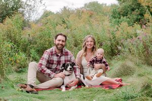 Smiling family with baby and black and white dog sitting on red blanket in lush garden meadow.