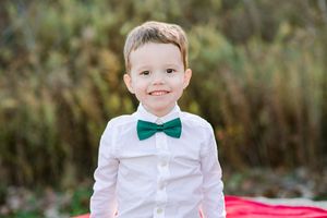 Adorable young boy in white shirt and green bow tie smiles during outdoor fall family photo session.