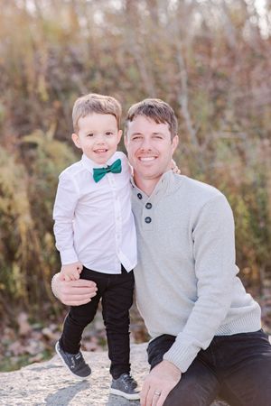 Smiling father crouches beside young boy in green bow tie and white shirt during outdoor fall portrait.