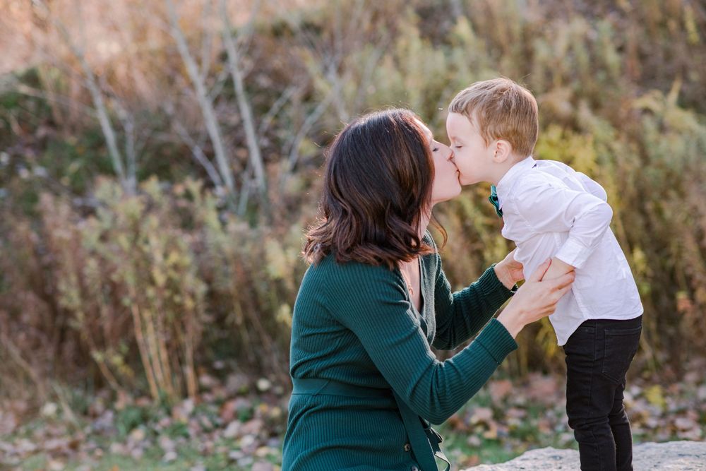 Loving mother kisses young boy in white shirt and green bow tie during outdoor fall family portraits.