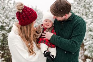 Parents in winter hats and sweaters smiling at baby in red plaid outfit in snowy pine forest.