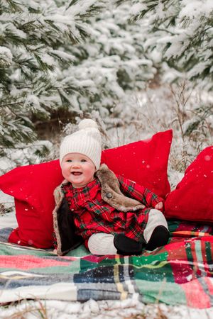 Happy baby in red plaid dress and white knit hat sitting on snowy blanket surrounded by red pillows.