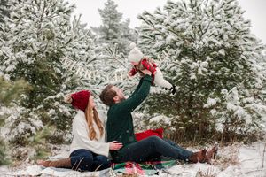 Couple sitting in snow lifting joyful baby in red plaid outfit with snow-covered pine trees behind.
