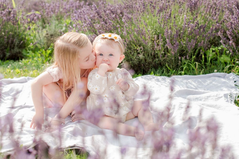 Sweet older sister kissing baby sibling on the cheek while sitting on a blanket in a lavender field.