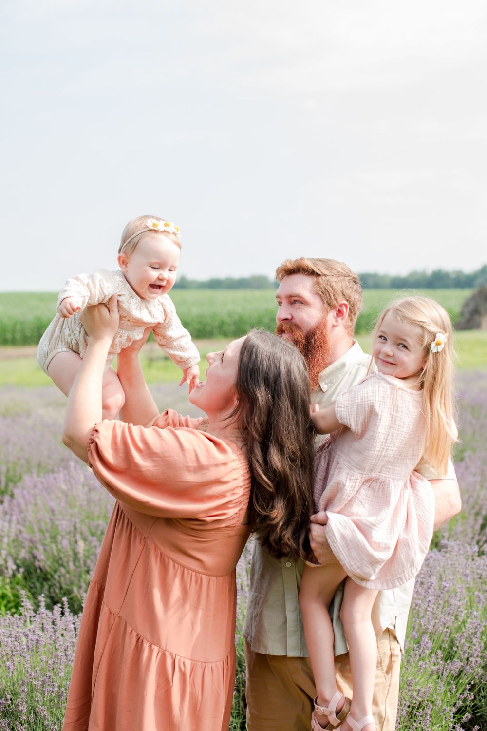Happy family in lavender field with mother lifting laughing baby and father holding smiling toddler girl.