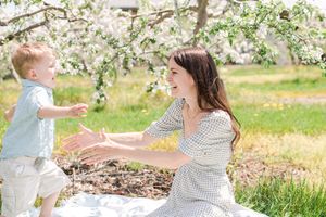 Mother reaching out to her toddler son under blooming white apple trees in spring.