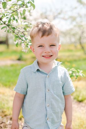 Smiling toddler boy in light blue shirt standing outdoors near blooming spring tree branches.