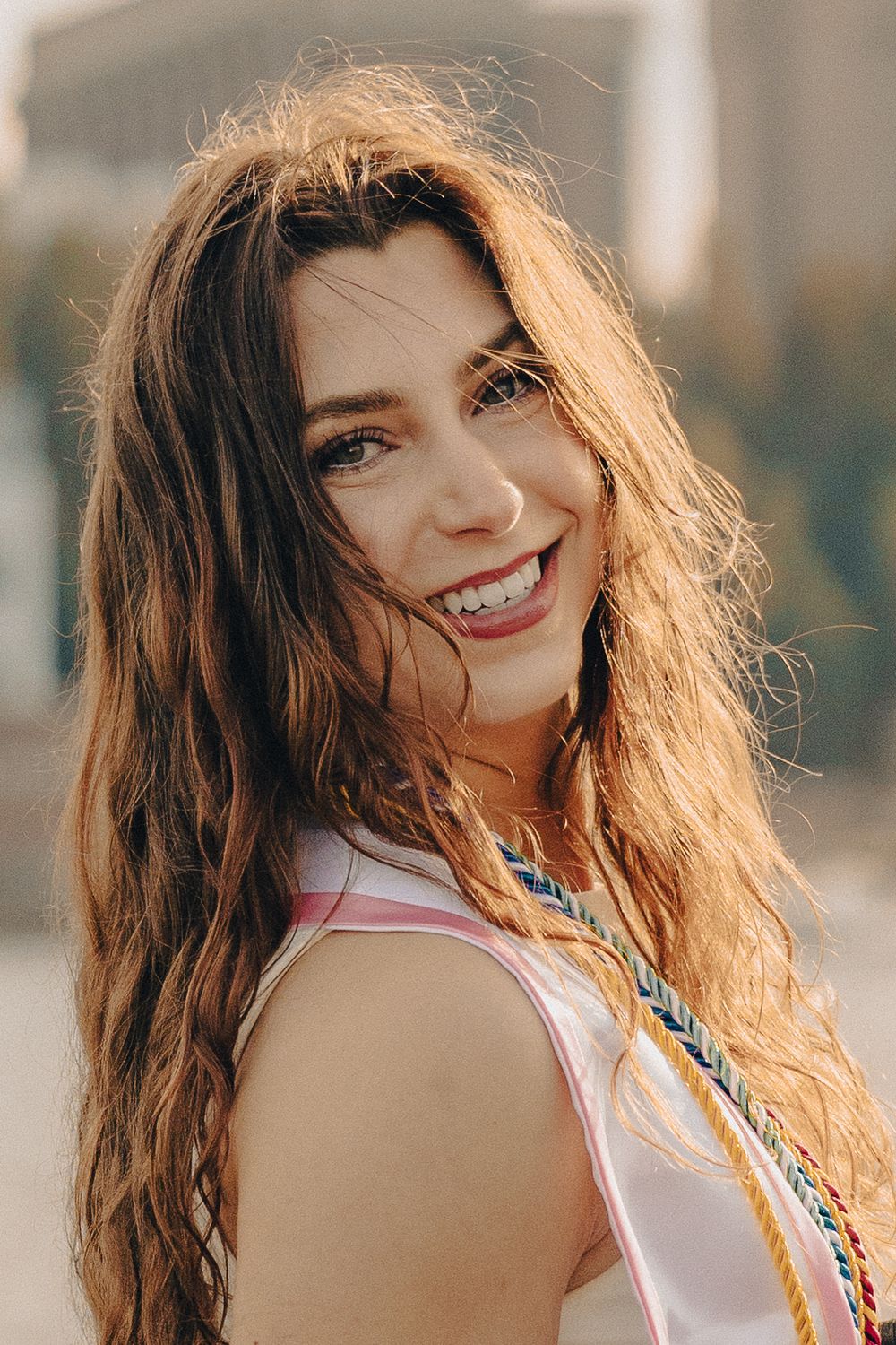 Smiling young woman with long wavy brown hair wearing a white top and colorful braided accessories outdoors.