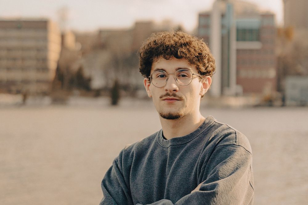 Young man with curly hair and glasses, arms crossed, posing outdoors in an urban setting during golden hour.