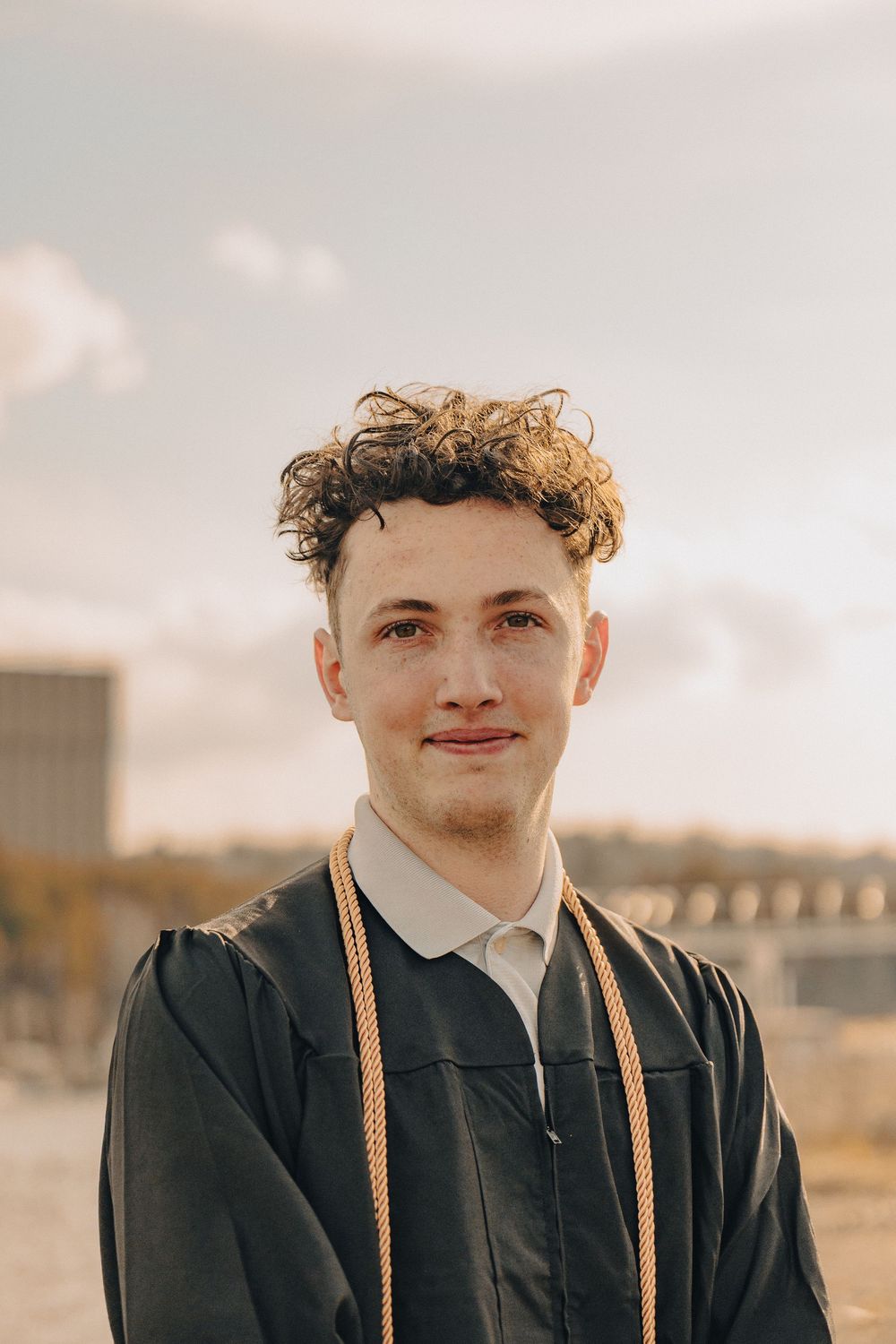 Young man in black graduation gown with gold honor cords smiling outdoors on a sunny day.