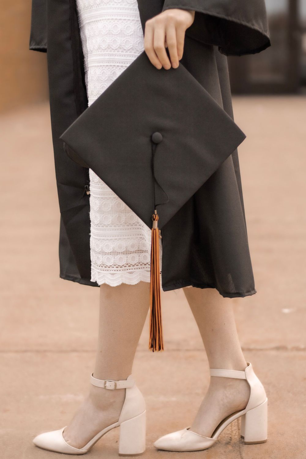 Close-up of graduation cap and white block heels against a black graduation gown and lace dress.