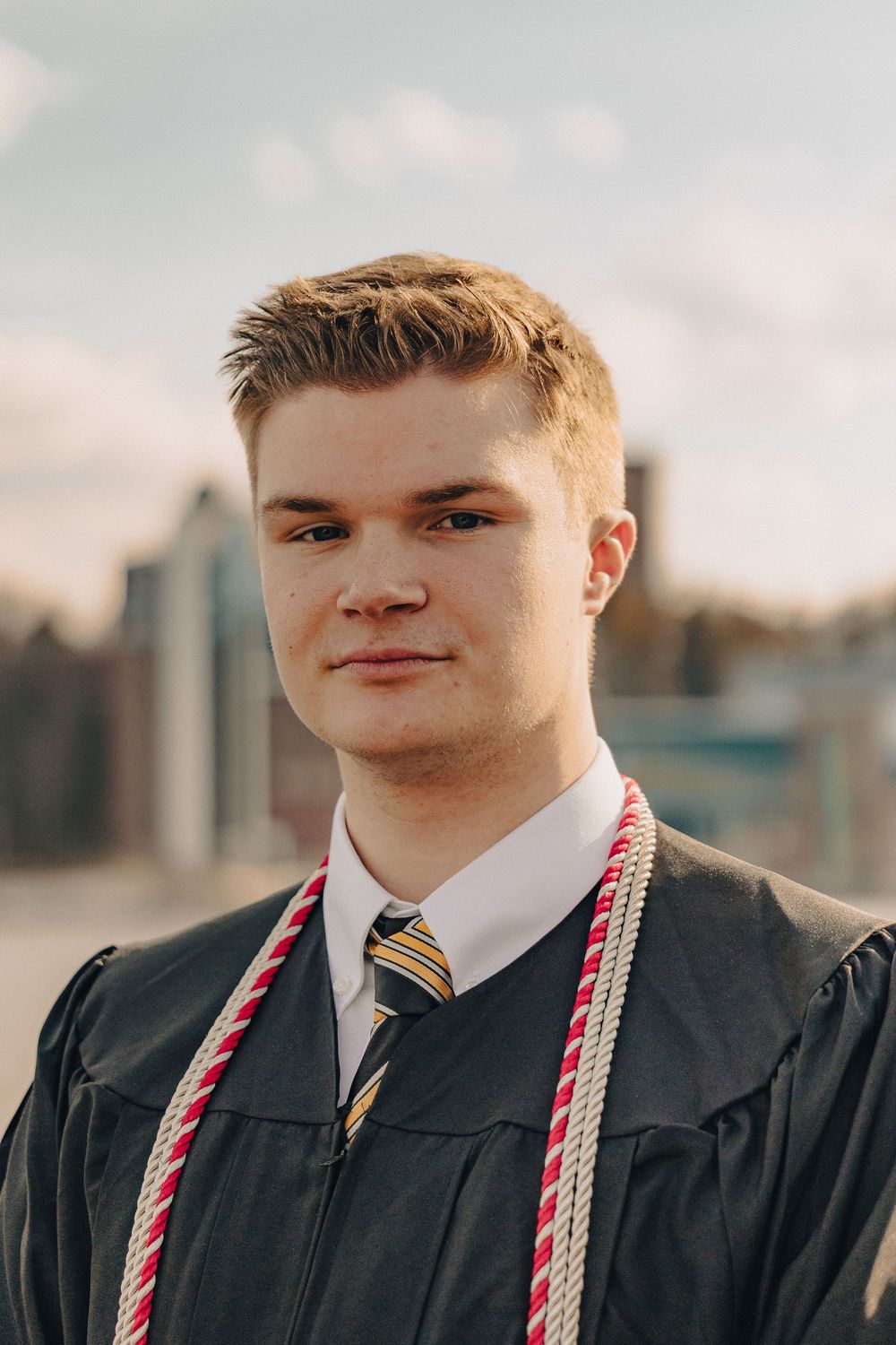 Young man in graduation gown and honor cords smiling outdoors during his graduation ceremony.