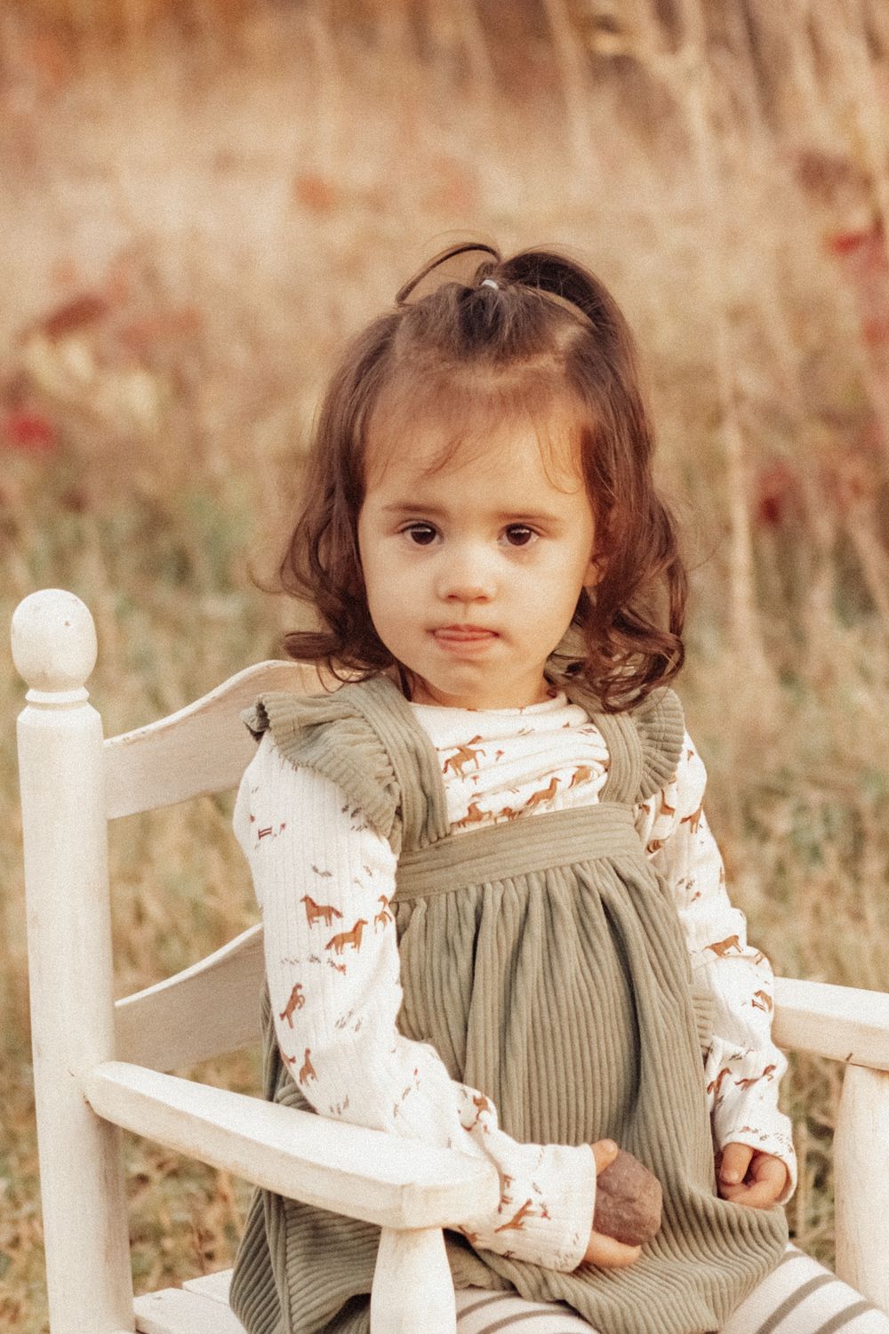 A small child in a gray jumper dress and floral top sits on a white wooden chair in an autumn field.