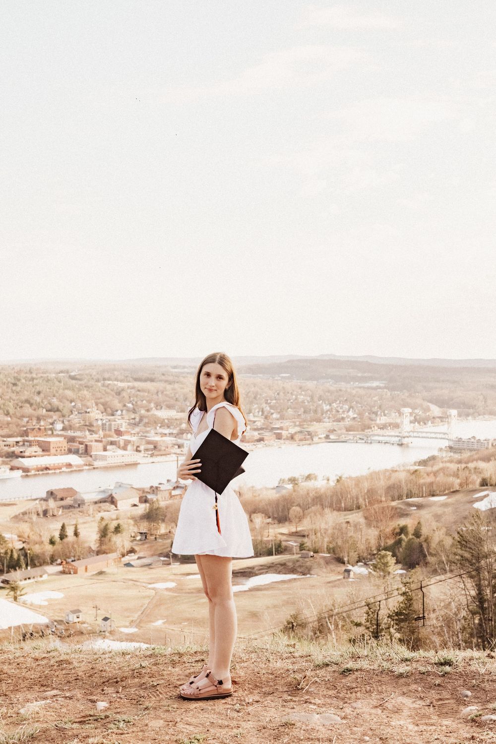 Graduate in white dress holding diploma on hilltop with scenic town view below.