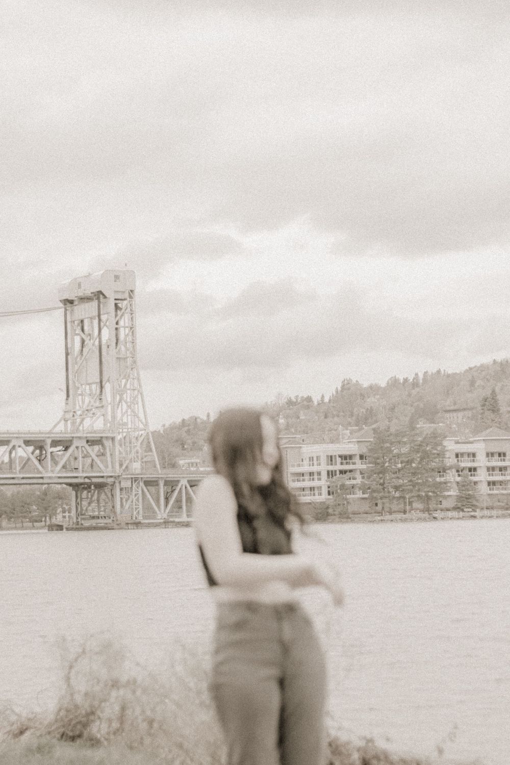 Artistic blurred photo of person by waterfront with bridge structure visible in background.