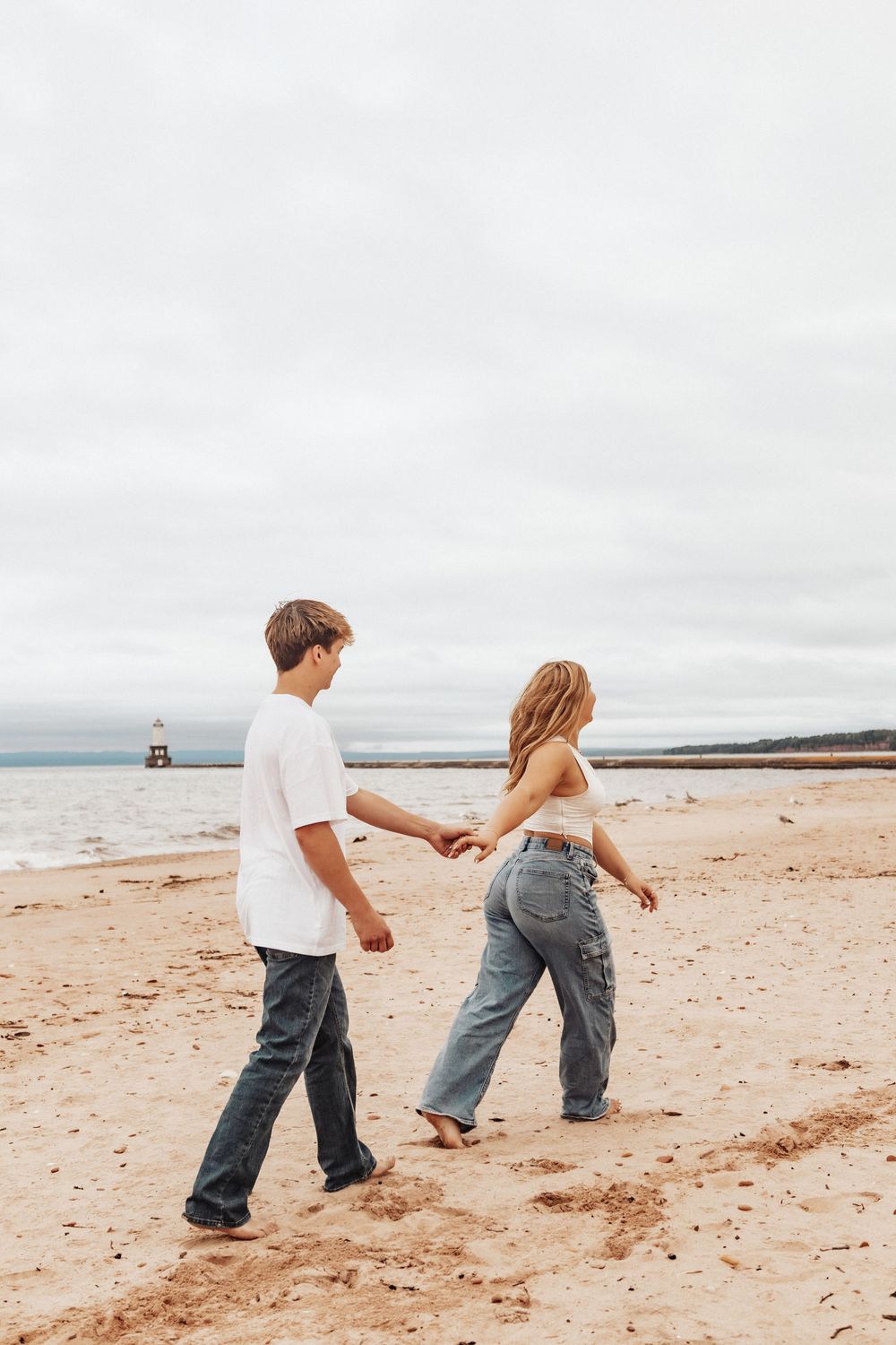 Young couple walking hand in hand on a beach with a lighthouse visible in the distance on an overcast day.