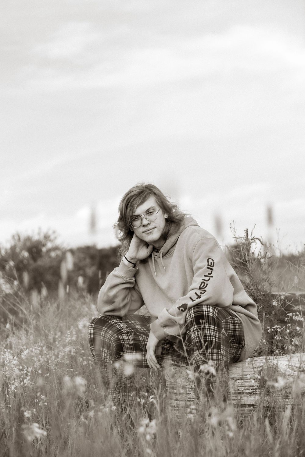 Black and white photo of a young person with long hair sitting in a wildflower field wearing a Carhartt hoodie and plaid pants.