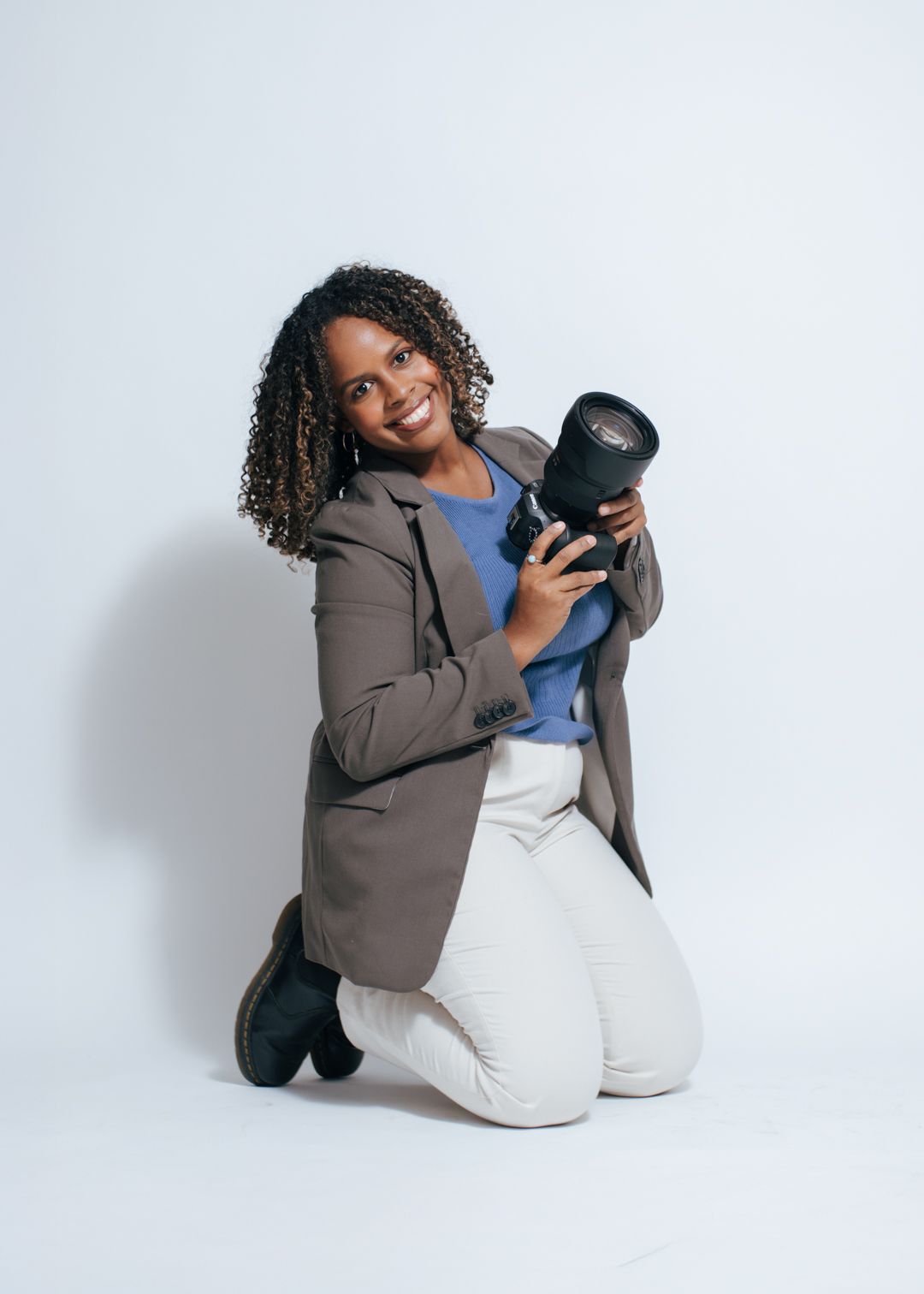 Professional photographer woman in gray blazer and white pants kneeling with camera lens against white background.