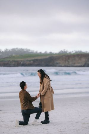 A couple shares an intimate moment during a proposal on a misty beach with cliffs in the background.