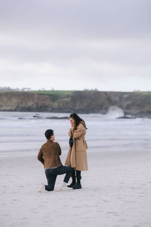 A surprise proposal moment on a sandy beach with rugged coastal cliffs visible in the distance.