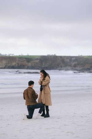 Two people in winter coats stand together on a foggy beach with coastal cliffs in the background.