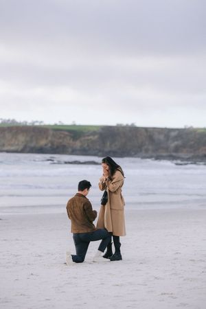 Two people share an intimate moment during a beach proposal with dramatic coastal cliffs behind them.
