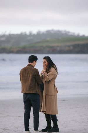 Two people share an emotional moment during a proposal on a cloudy beach with coastal cliffs behind them.