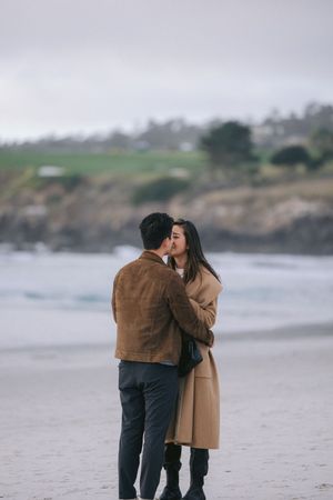 A couple shares a tender embrace on a misty beach with rocky cliffs and ocean waves in the background.