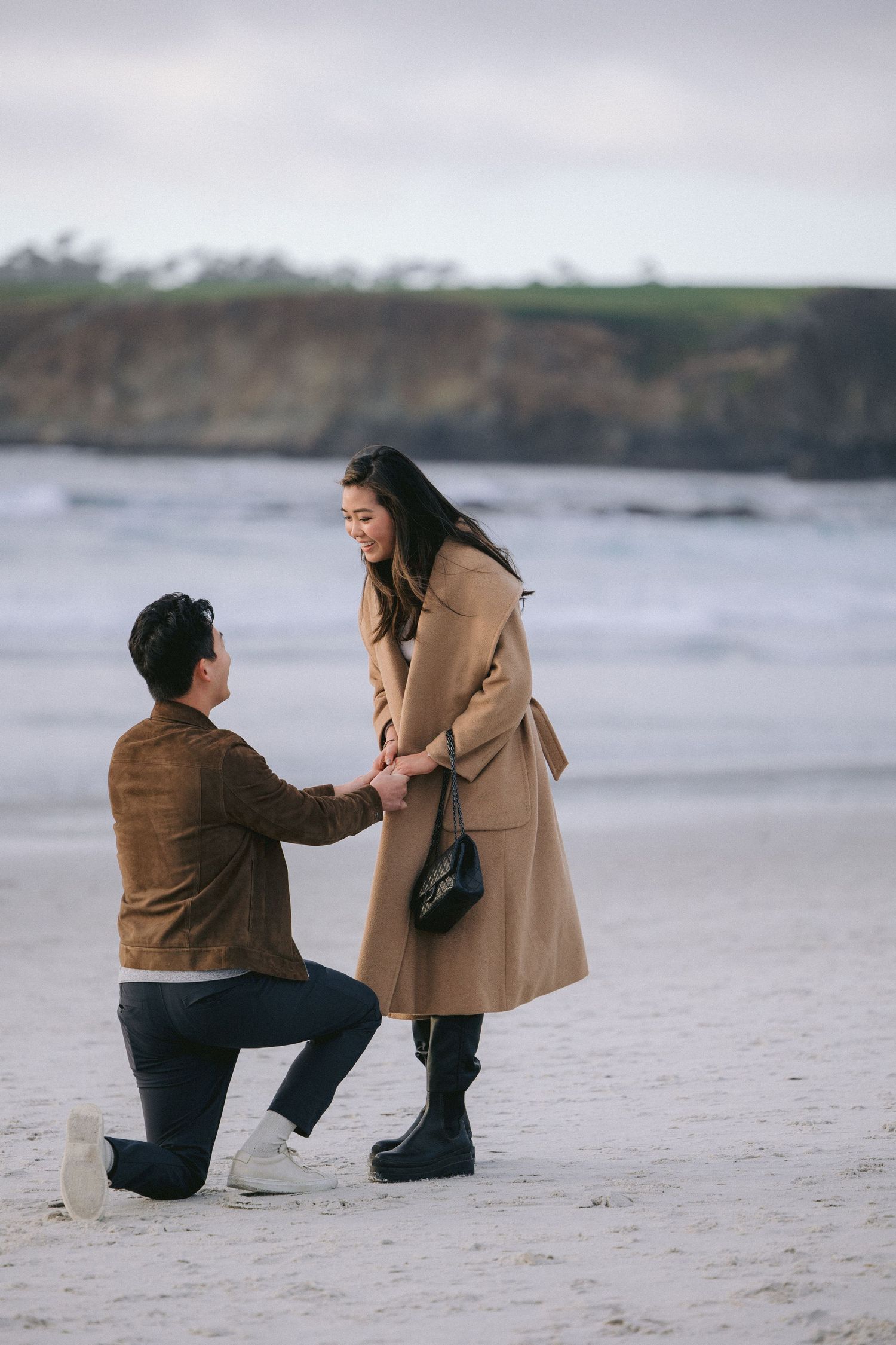 A romantic beach proposal scene with two people embracing near ocean cliffs on a cloudy day.