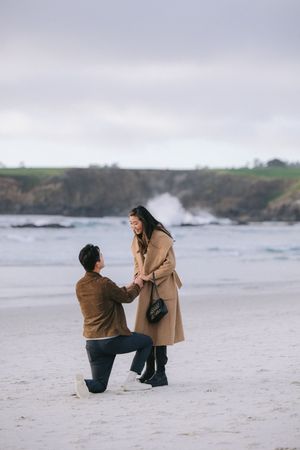A romantic beach proposal moment captured against dramatic coastal cliffs and crashing waves on a moody day.