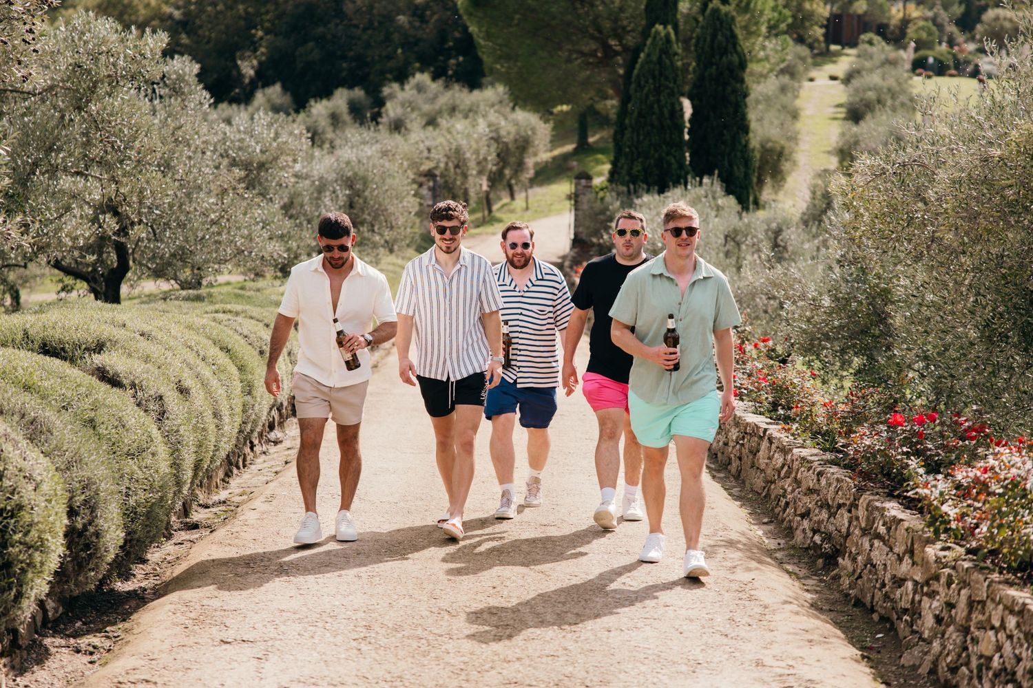 Friends walking together on a stone path surrounded by olive trees in a Mediterranean landscape.