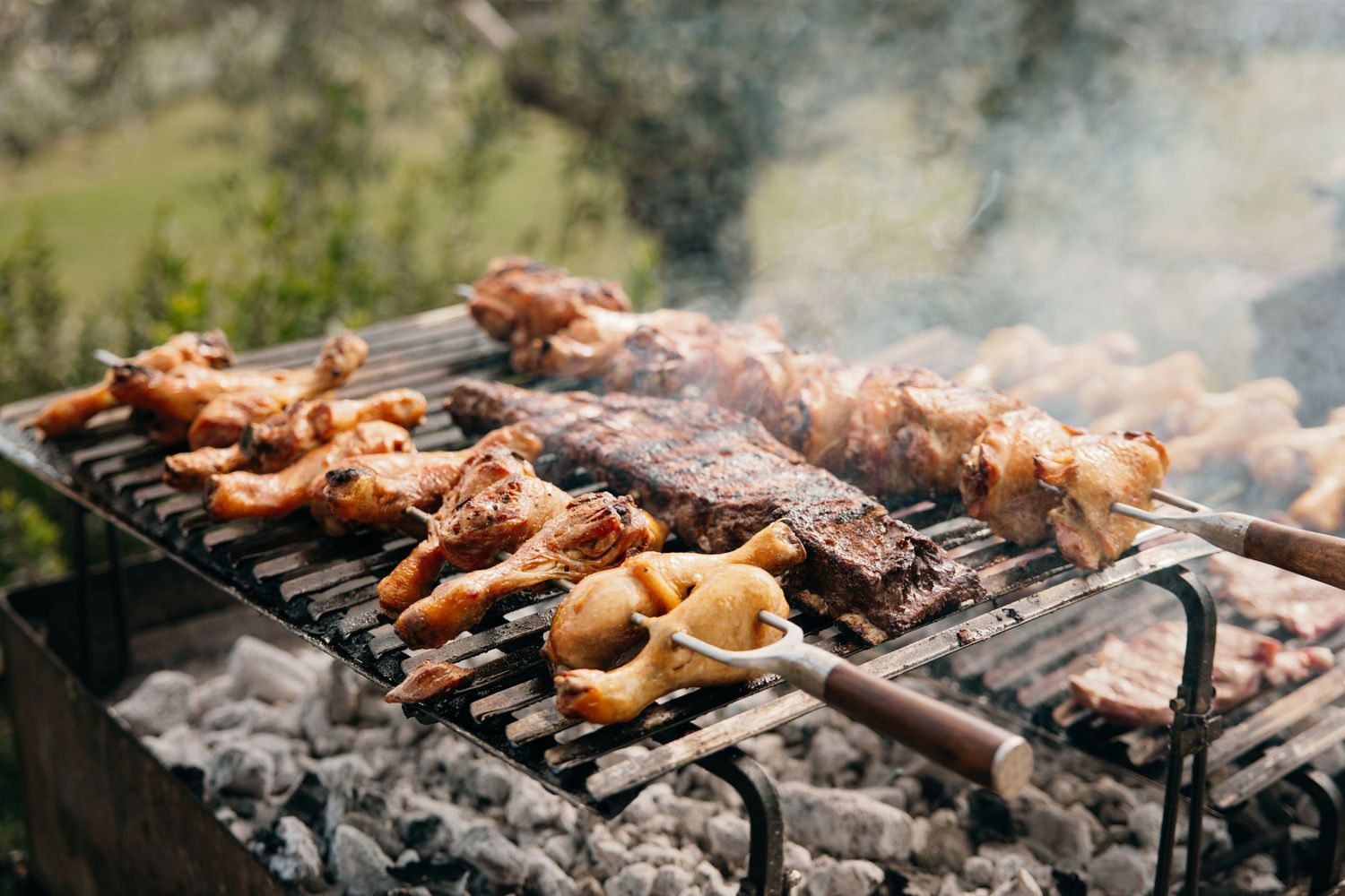 Various meats grilling over charcoal with smoke rising in an outdoor setting.