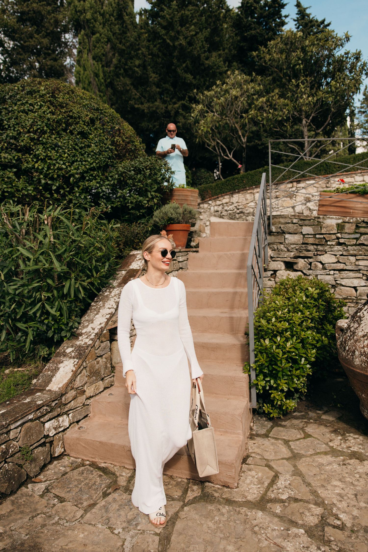 Woman in white flowing dress descends stone steps at a scenic garden venue.